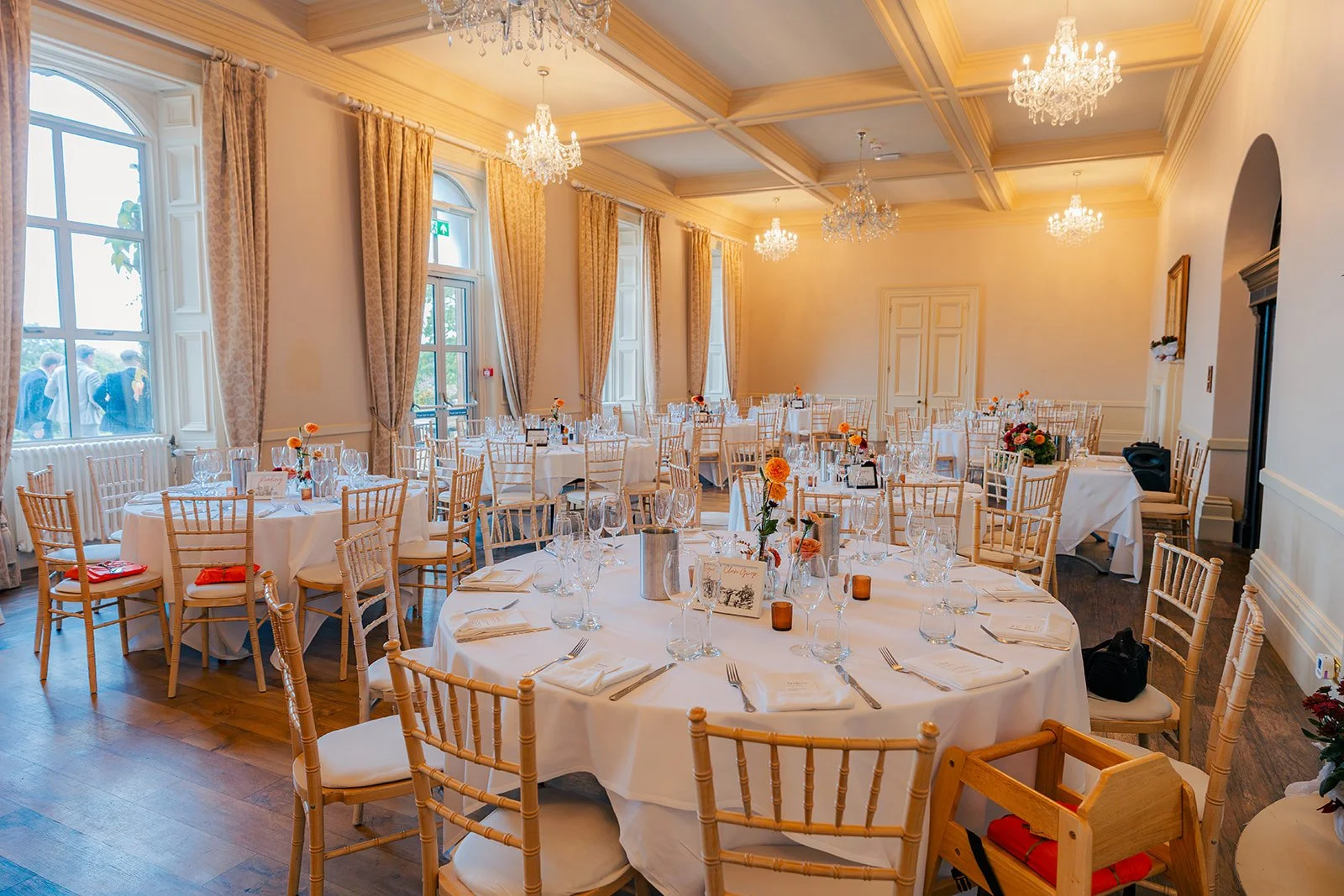 Wedding breakfast room at Eastwood Park in Gloucestershire, with round tables, gold chairs, chandeliers and orange floral details, photographed by Just James Weddings for elegant venue inspiration.