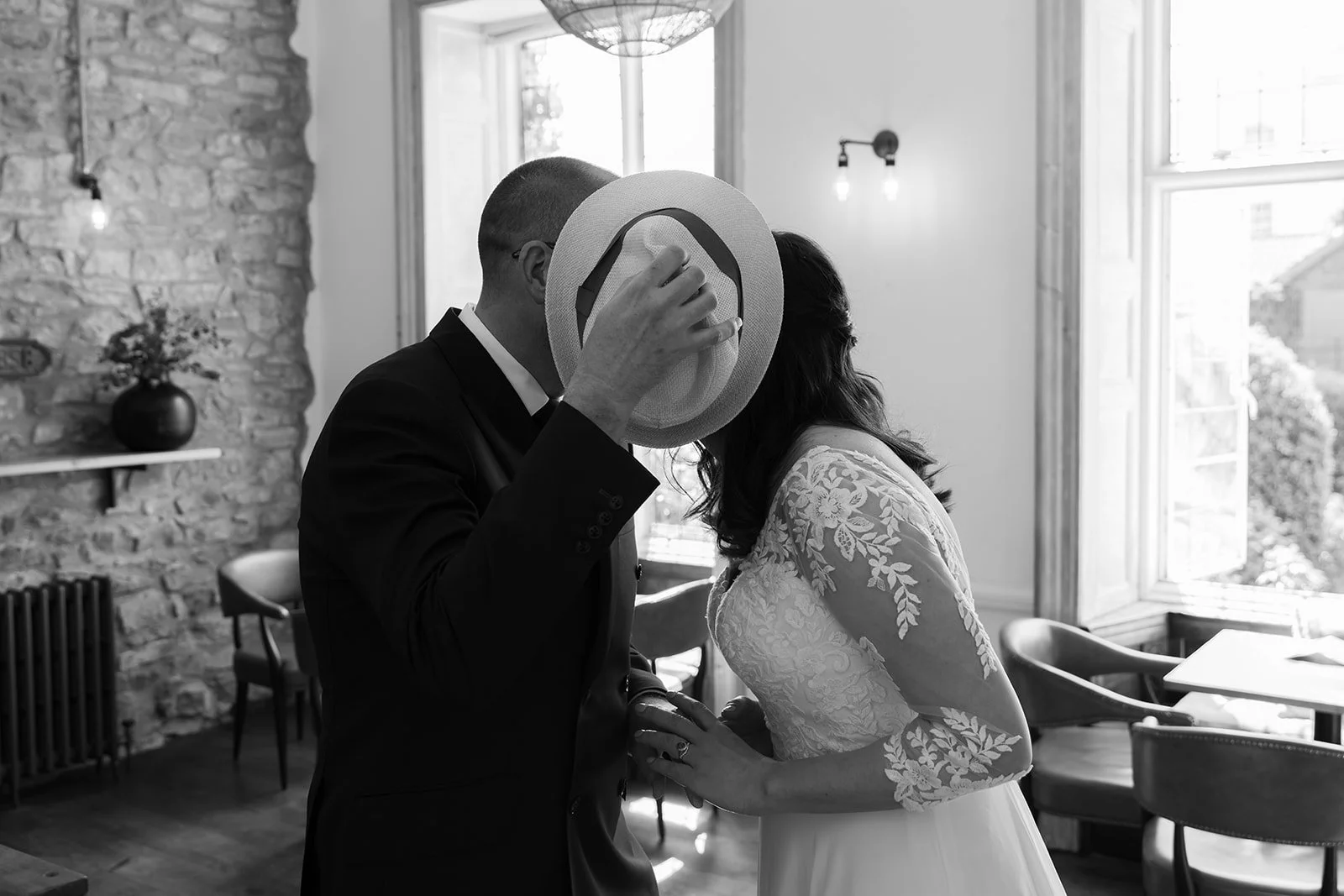 Black and white candid photo of Zoe and Paul sharing a kiss behind Paul’s hat during their wedding at No. 4 Clifton Village in Bristol, UK, capturing a playful private moment.