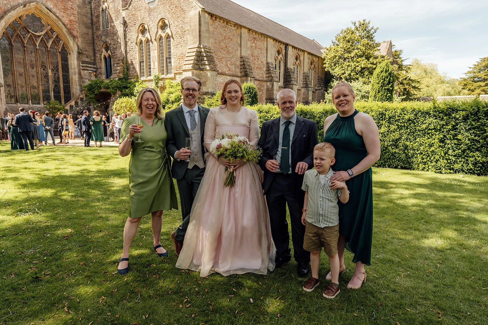 Charlotte & Jack family portrait at Bishop’s Palace in Somerset, UK, with the couple standing on the lawn beside close family and a child, holding drinks and flowers as guests mingle near the historic palace buildings behind them in sunshine.