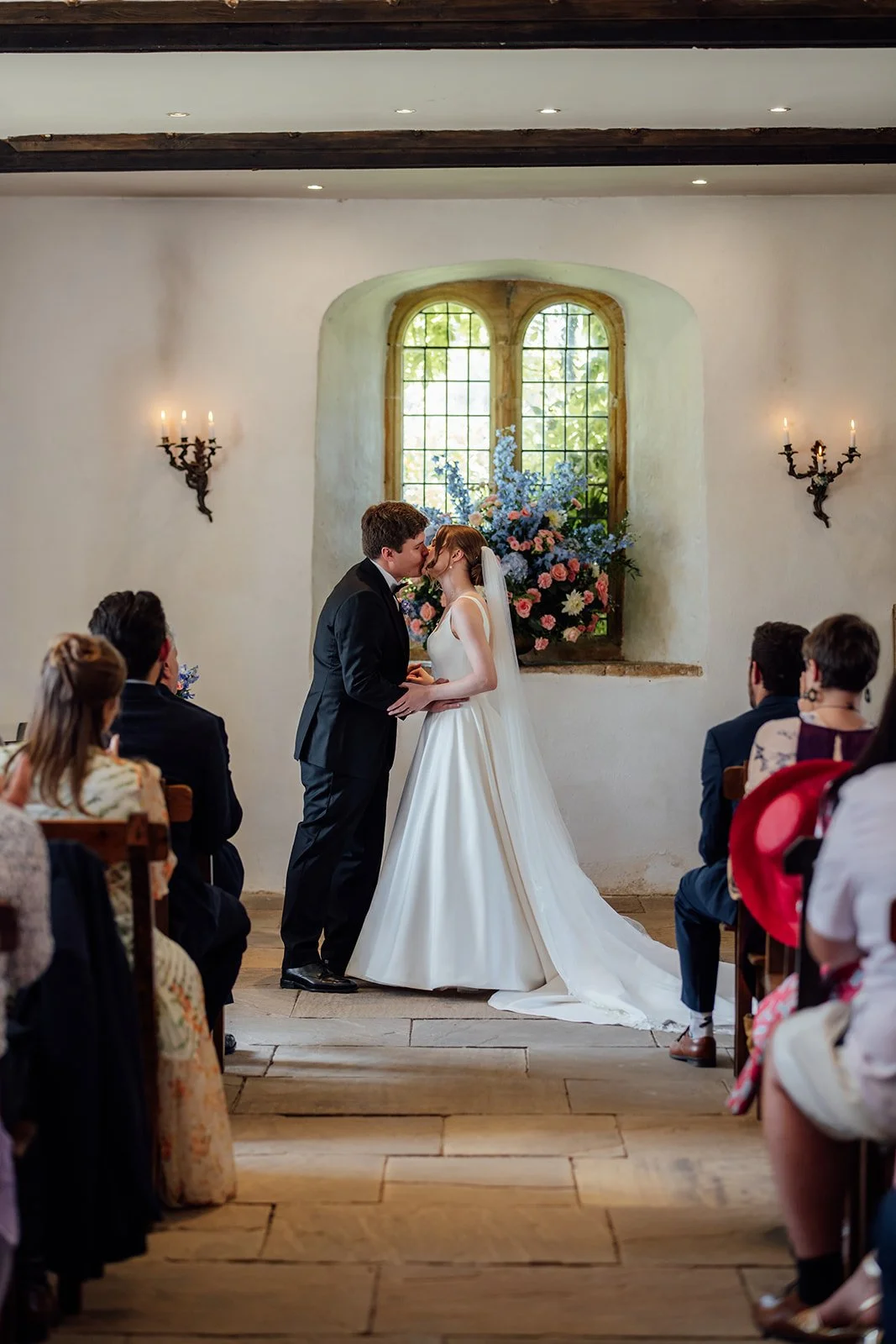 Rachel & Tom kiss during their indoor wedding ceremony at Brympton House in Somerset, framed by a large colourful floral arrangement, photographed by Just James Weddings.