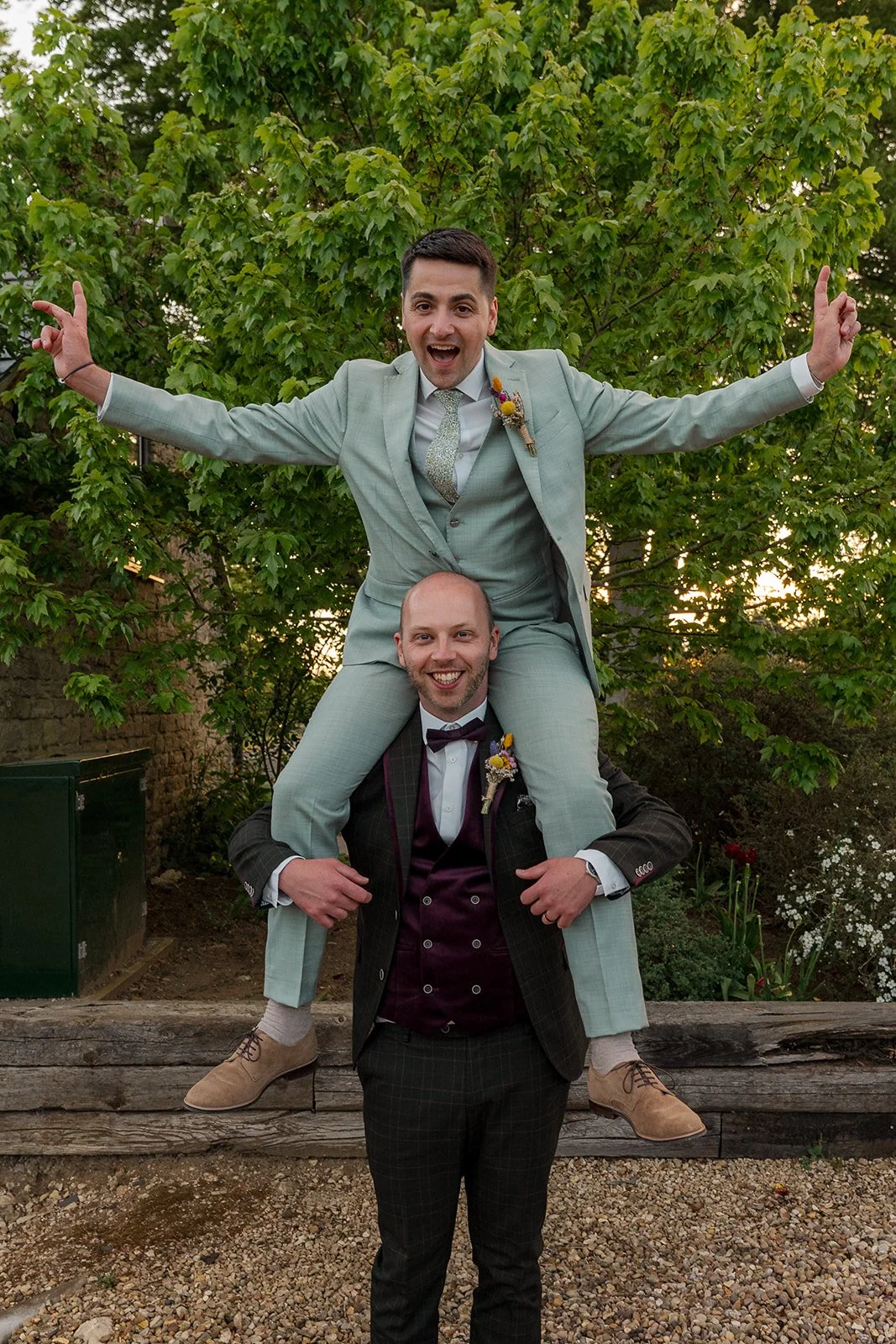 Sam perches on Gavin's shoulders with his arms thrown wide in celebration during their wedding day at The Great Barn in Somerset, both grooms beaming in their coordinating suits with floral buttonholes, surrounded by lush green trees.
