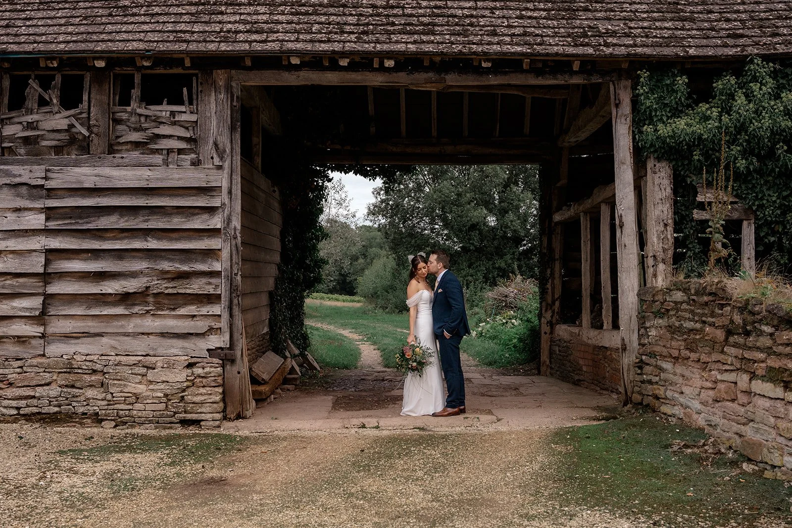 Lilly & Lloyd kissing beneath a rustic timber archway at Broadfield Court in Herefordshire, photographed by Just James Weddings for romantic countryside wedding portraits.
