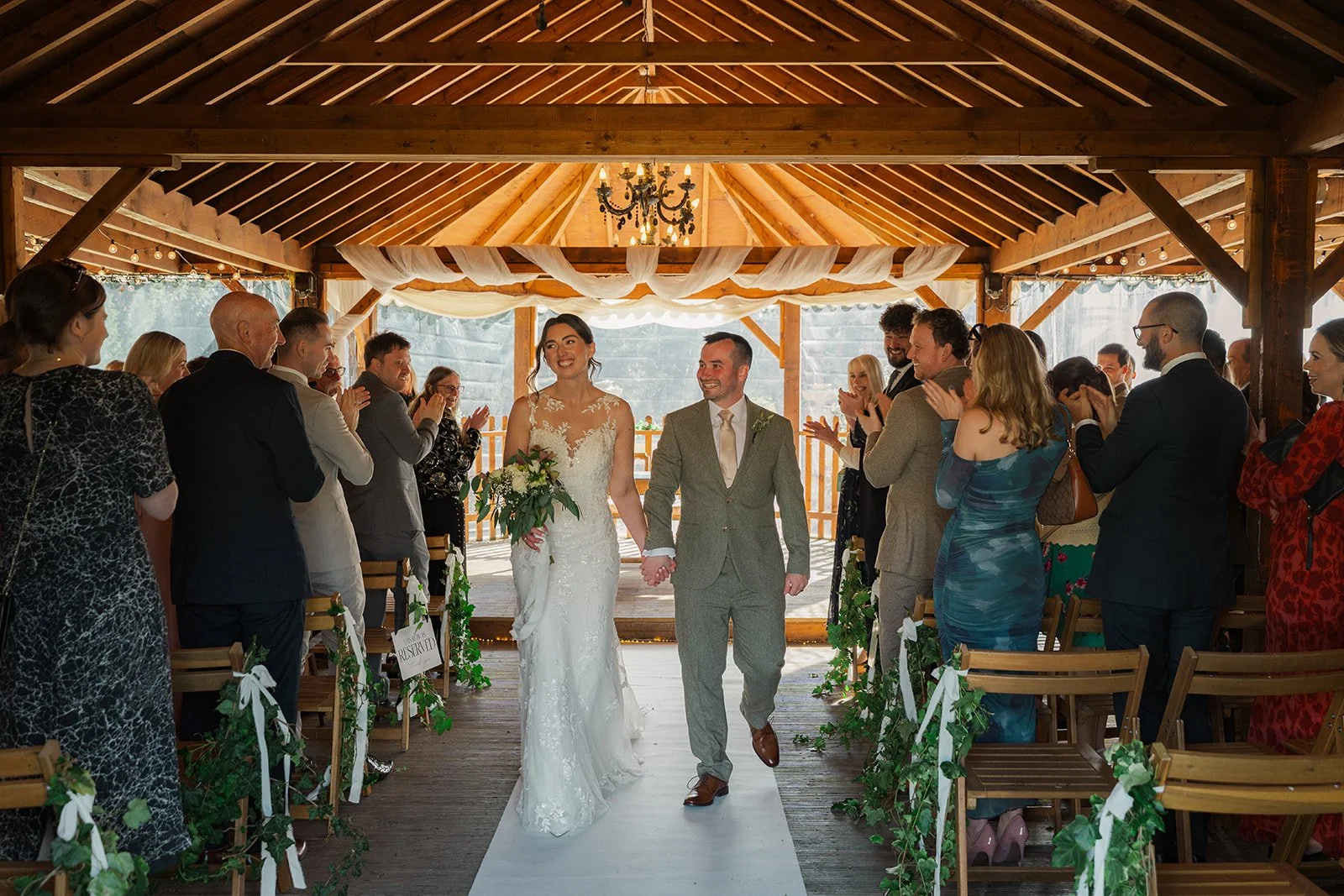 Anna & Chris walk back down the aisle at Elmhay Park in Somerset, UK, smiling as guests stand and applaud inside the timber ceremony barn. A joyful just-married moment from Anna & Chris’s wedding, full of warmth, celebration, and natural elegance.