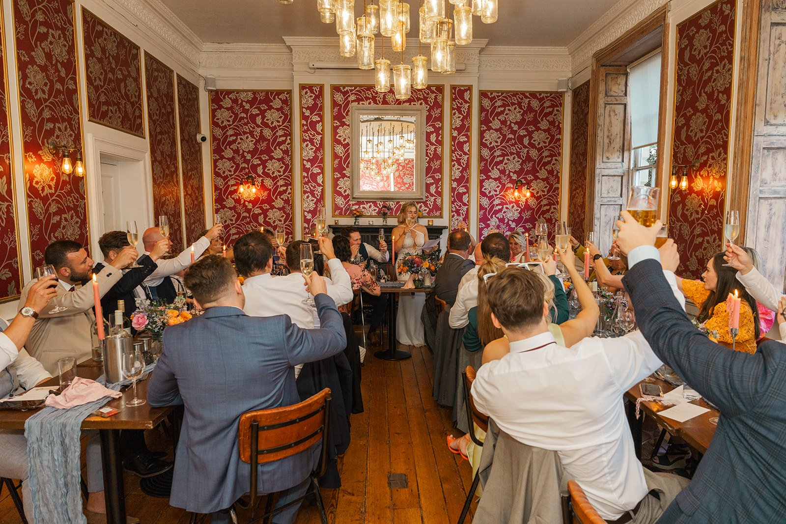 Wedding guests raise their glasses for a toast during Kristy and Will’s reception at No. 4 Clifton Village in Bristol, UK, inside the venue’s elegant red and gold dining room.
