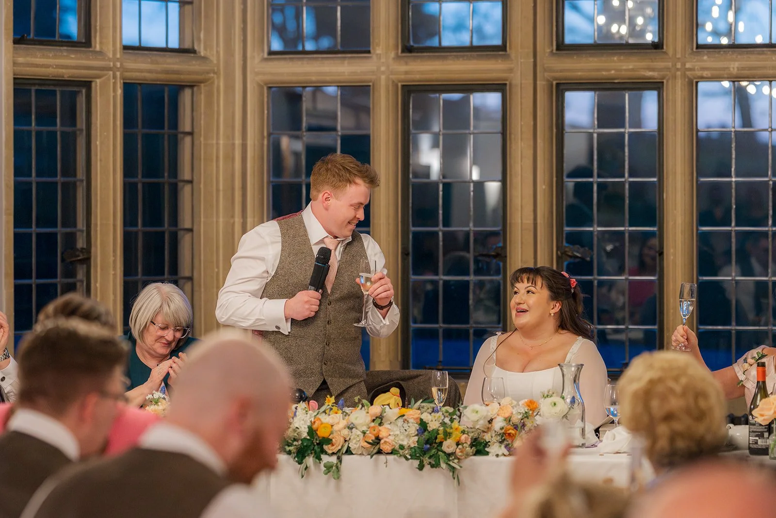 Josh gives a wedding speech beside Emily during their reception at Coombe Lodge in Somerset, UK, surrounded by guests and colourful floral arrangements.