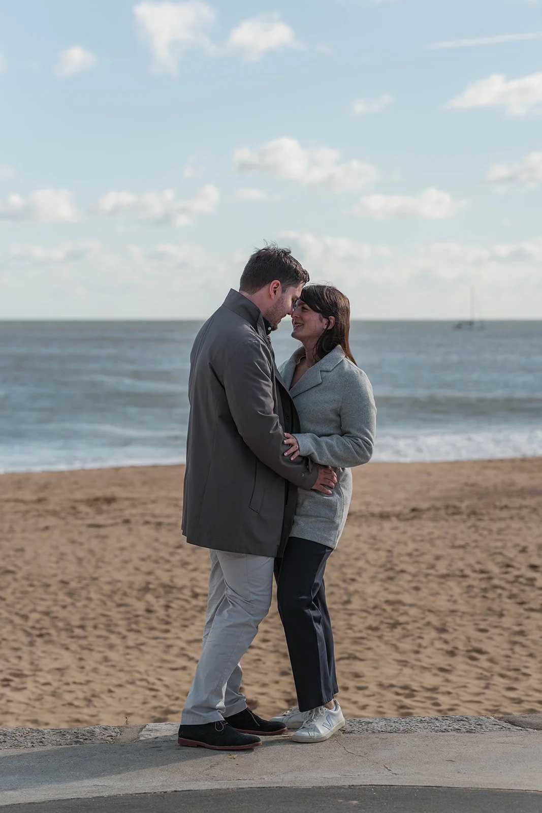 Valentina & Nicky stand close during their engagement shoot on the beach promenade, smiling with their foreheads together as the sea and soft clouds sit behind them. Their relaxed pose and gentle expressions create an intimate coastal portrait.