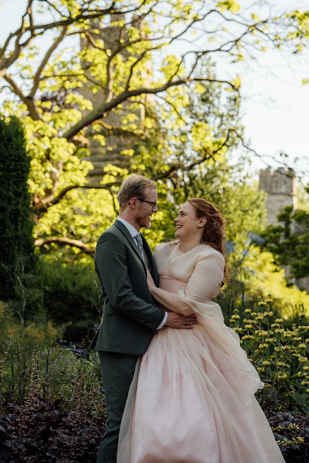 Charlotte & Jack couple portrait at Bishop’s Palace in Somerset, UK, with the bride and groom laughing together in the gardens, surrounded by soft flowers, greenery and golden light after their ceremony in the historic Wells city wedding venue.