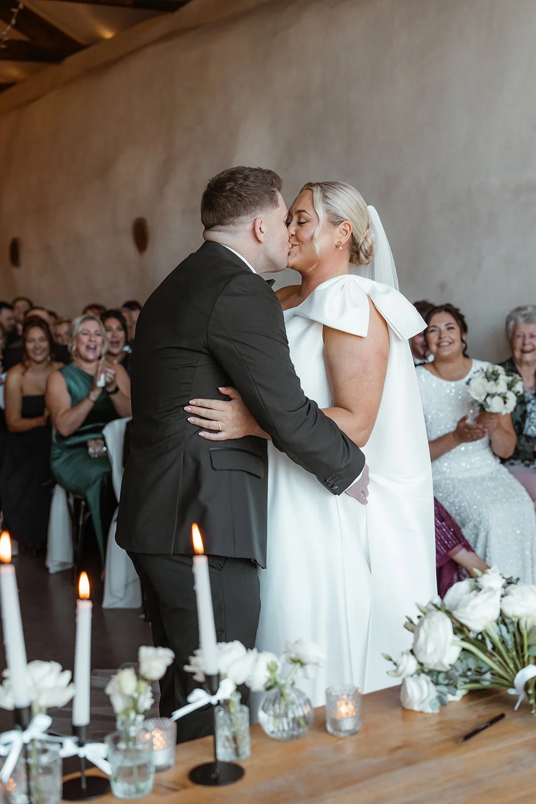 Jessie & Jay share their first kiss at Upton Barn & Walled Garden in Devon, UK, during a candlelit indoor wedding ceremony, surrounded by smiling guests and soft florals, with the bride and groom framed in a warm, elegant barn setting indoors.