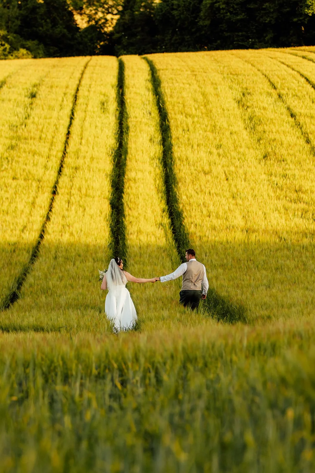 Chloe & Armani walking hand in hand through a golden field at The Barn at Upcote in the Cotswolds, photographed by Just James Weddings for relaxed countryside wedding photography.