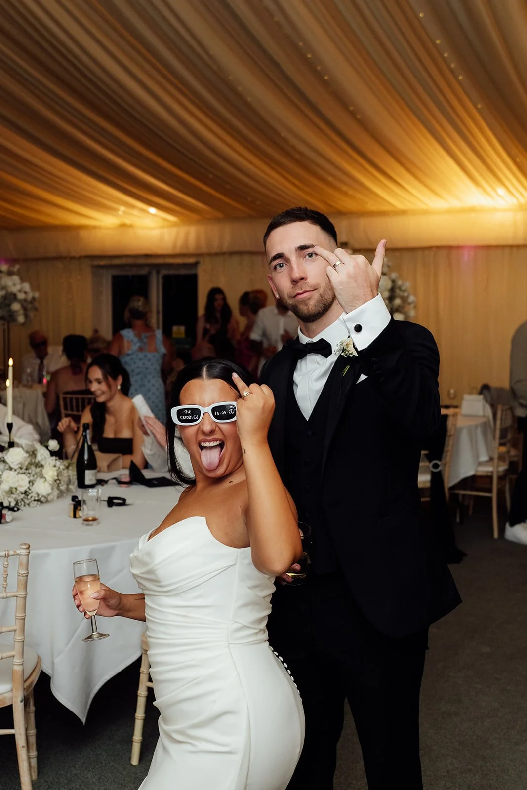 Nisha and Jack pose playfully during their wedding reception at Batch Country House in Somerset, UK, with Nisha wearing white novelty sunglasses and holding a glass while both show off their wedding rings, photographed by Just James Weddings.