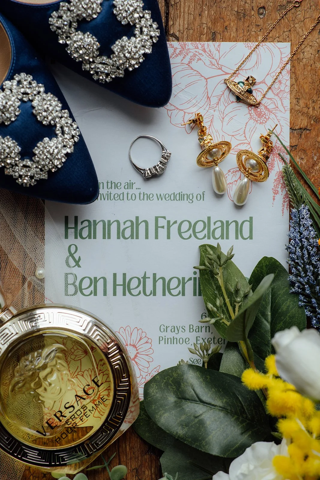Flat lay of Hannah & Ben’s wedding details at Grays Barn in Devon, UK, featuring navy embellished bridal shoes, perfume, earrings, ring, invitation stationery and colourful flowers on rustic wood, photographed beautifully by Just James Weddings.