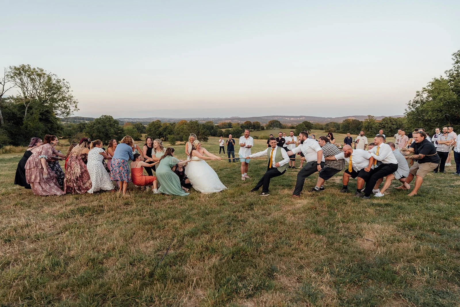 Lucy and Matthew joining their wedding guests for a lively outdoor tug of war in the Somerset countryside at Curated Canvas at Noah’s Pond, photographed by Just James Weddings.