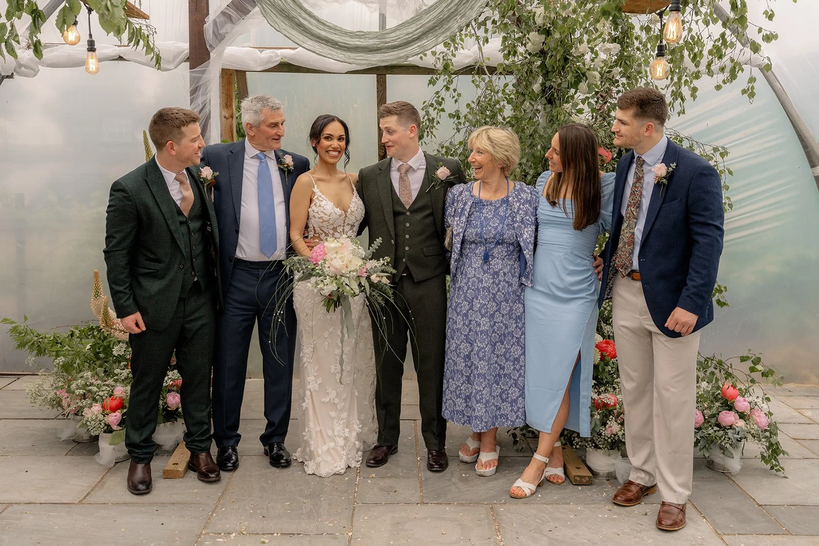 Jaye & Josh stand with family for a joyful group portrait at Farrington's Farm in Bristol, surrounded by soft greenery, pink flowers, draped fabric, pale blue outfits and warm festoon bulbs inside the relaxed poly tunnel wedding ceremony space.