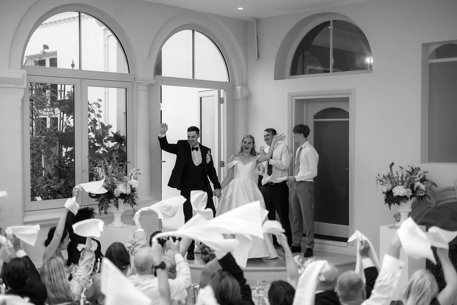 Black and white photo of Caitlin and Kevin entering their wedding reception at Clevedon Hall in Somerset, waving to guests as white napkins are raised in celebration.