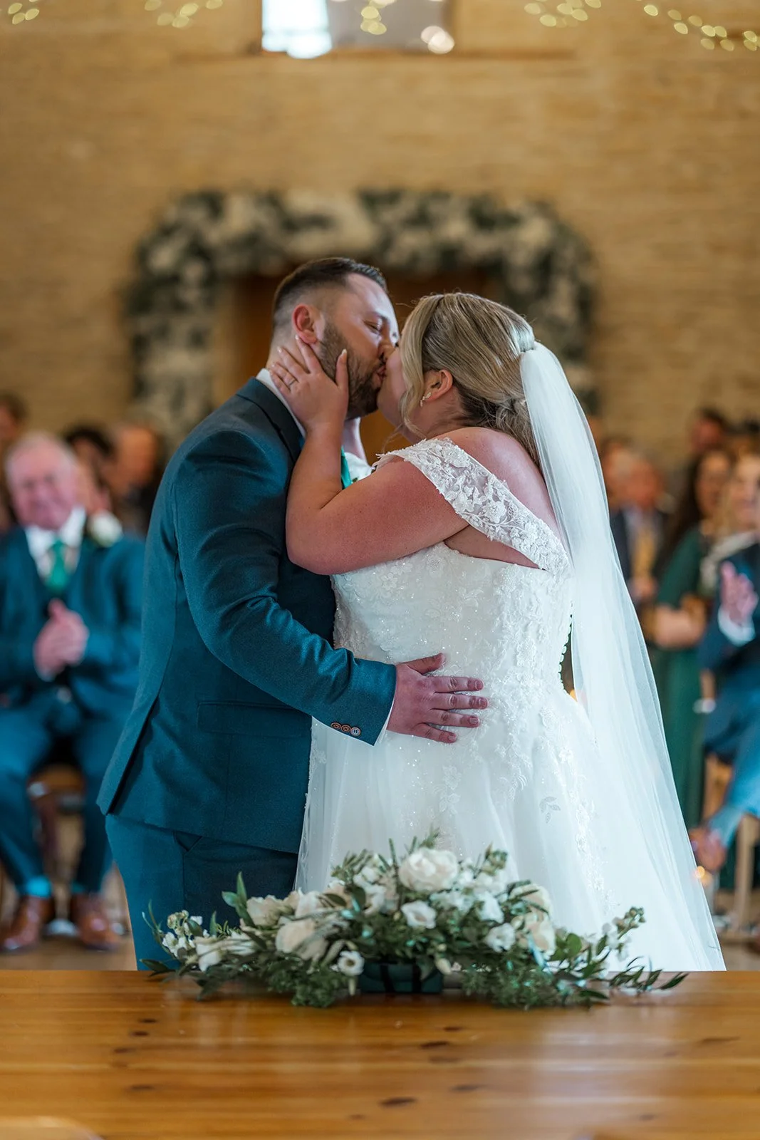 Siobhan & Nicholas share their first kiss during their wedding ceremony at Kingscote Barn in the Cotswolds, UK, surrounded by guests and soft floral details, a romantic rustic barn wedding moment captured at the altar as family and friends watch on.