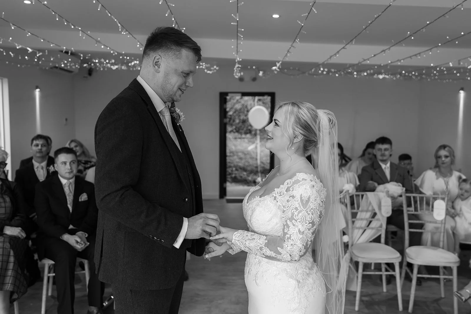 Daisy & Andrew holding hands during their indoor ceremony at Sandy Cove Hotel in North Devon, surrounded by guests and fairy lights, captured in black and white by Just James Weddings for emotional coastal wedding ceremony photography moments.