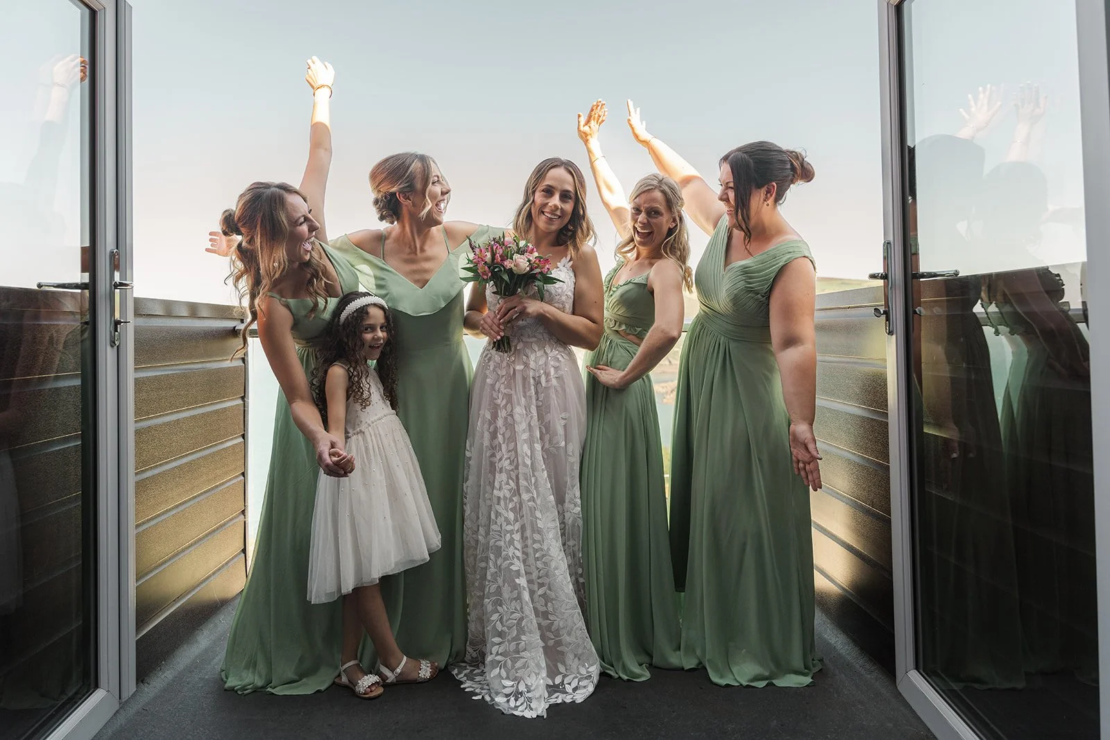 Toya & Simon celebrating with bridesmaids and a young flower girl on the balcony at Sandy Cove Hotel in North Devon, with sea views behind them, photographed by Just James Weddings for joyful coastal wedding party portraits and inspiration.