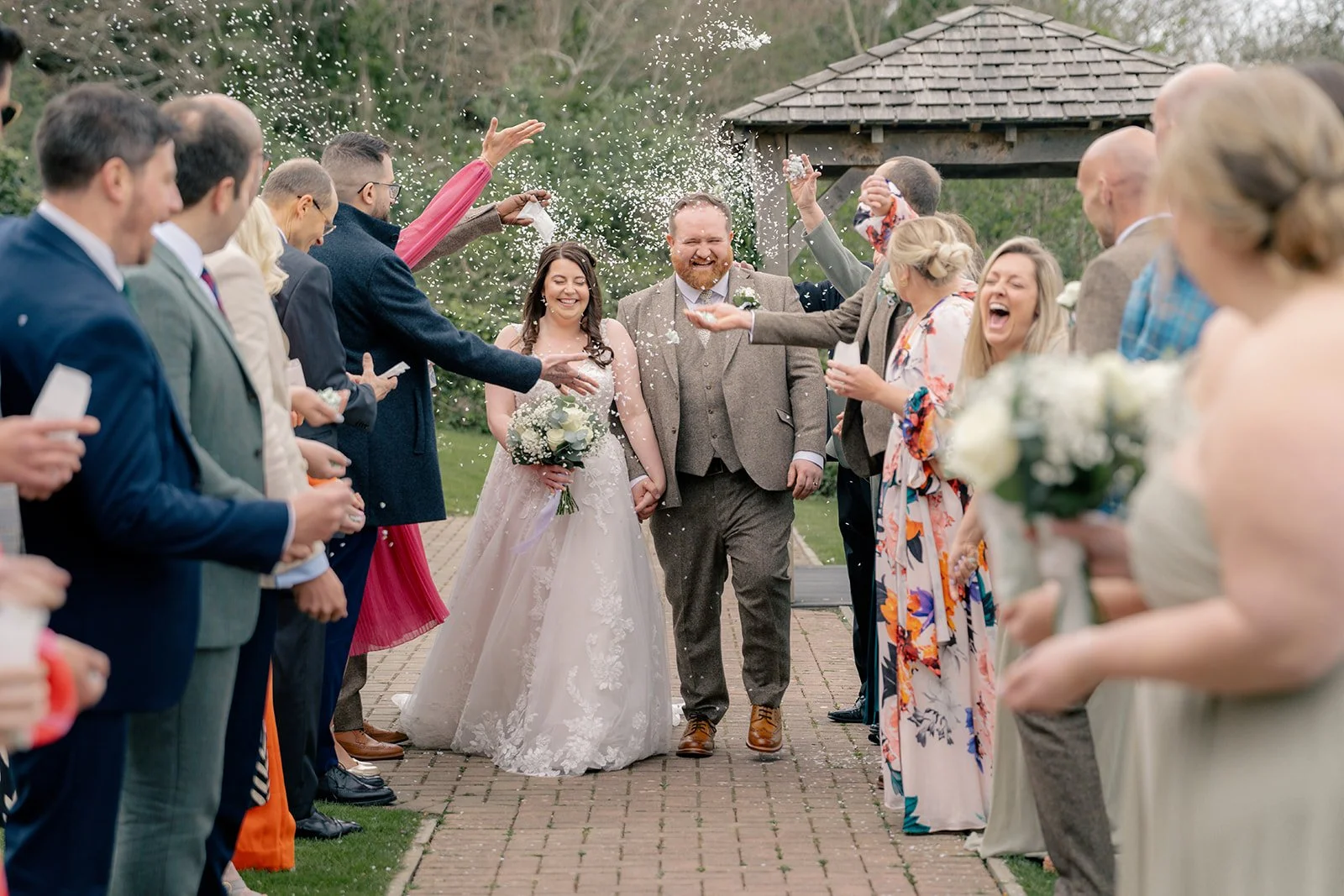Sam & Terri walking through a joyful confetti line at Sandy Cove Hotel in North Devon, smiling with guests gathered on both sides, photographed by Just James Weddings in a relaxed documentary wedding photography style after their ceremony vows.