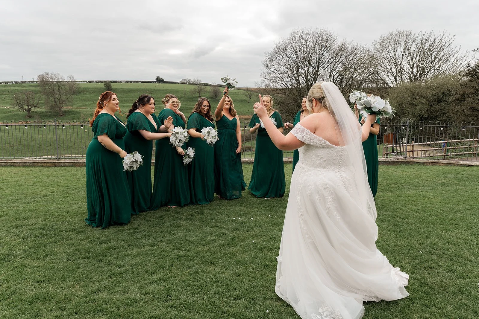 Siobhan throws her bouquet to bridesmaids in emerald green dresses on the lawn at Kingscote Barn in the Cotswolds, UK, during Siobhan & Nicholas' wedding, a fun outdoor barn wedding moment with countryside views and bridal party reactions.