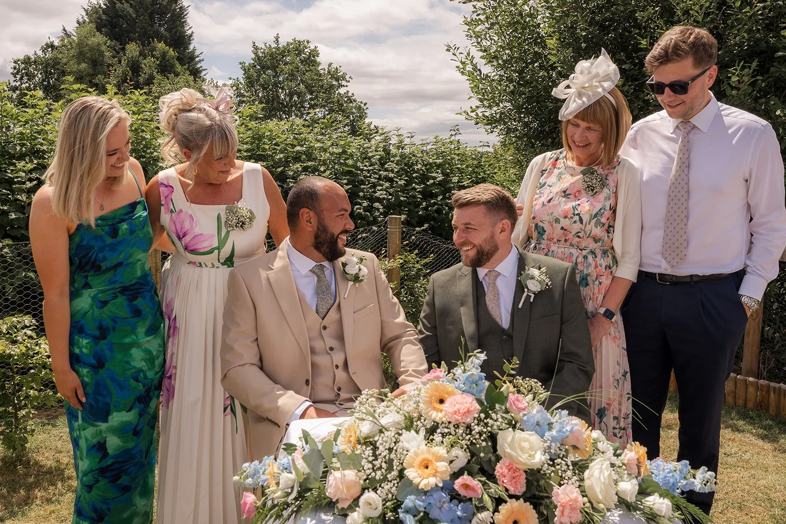 Aaron & Jack smile together with close family members behind a colourful floral arrangement after their outdoor ceremony at Rock Village Hall in Worcestershire, photographed by Just James Weddings.