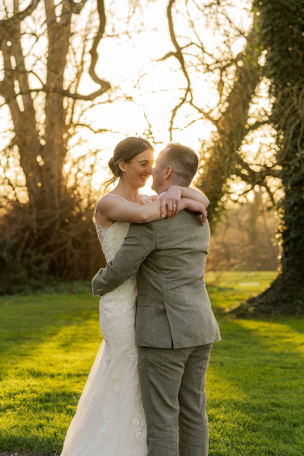 Anna and Chris embrace in golden evening light at Elmhay Park in Somerset, UK, smiling closely beneath the trees as the sun shines through behind them. A warm, intimate portrait from Anna & Chris’s wedding day, full of joy, connection, and softness.