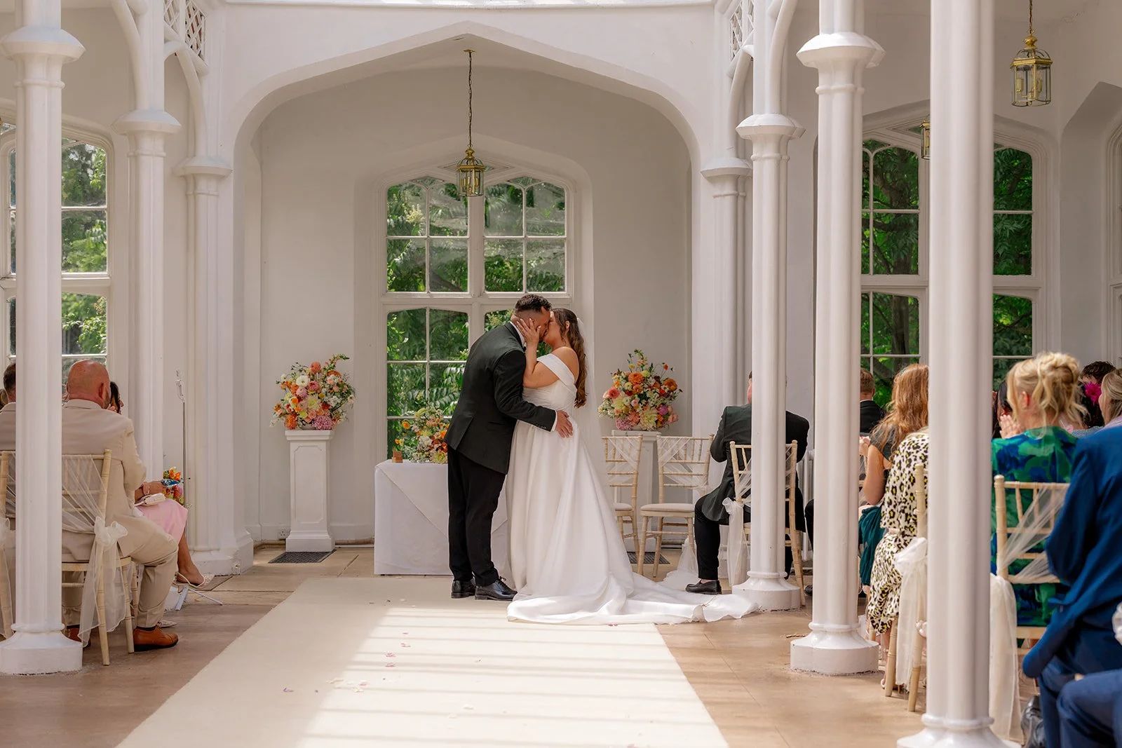 Leacarla & Carl share their first kiss as newlyweds during an elegant indoor ceremony at St. Audries Park in Somerset, UK, framed by white arches, floral arrangements, and seated guests as natural light fills the historic venue.