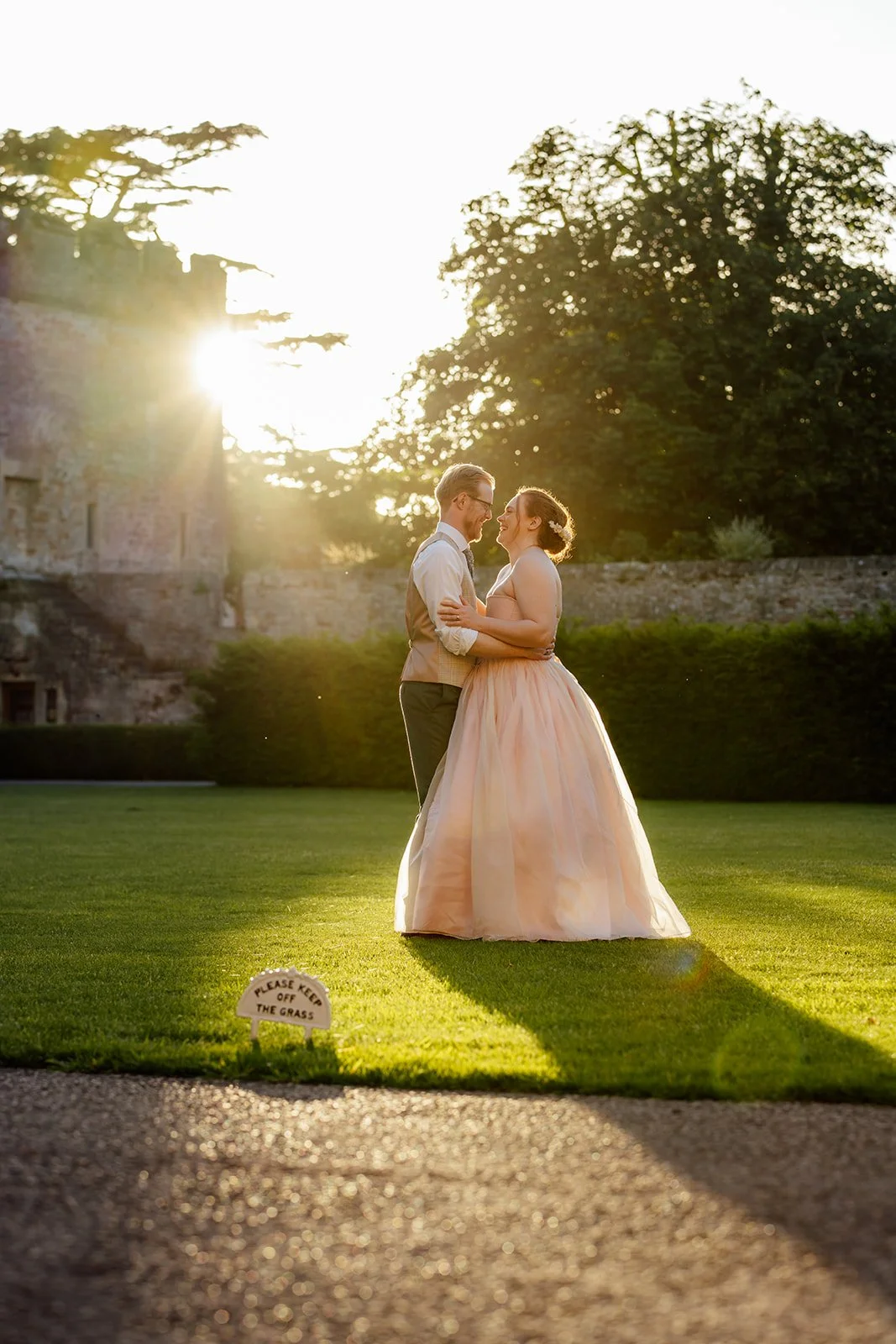 Charlotte & Jack stand together on the lawn at Bishop’s Palace in Somerset, UK, laughing in golden evening light beside the old stone walls, with the bride in her pink dress and the groom in shirtsleeves during their sunset couple portraits.
