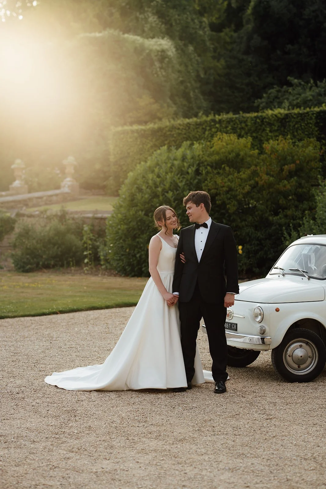 Rachel & Tom stand together beside a vintage white car in the evening light at Brympton House in Somerset, photographed by Just James Weddings.