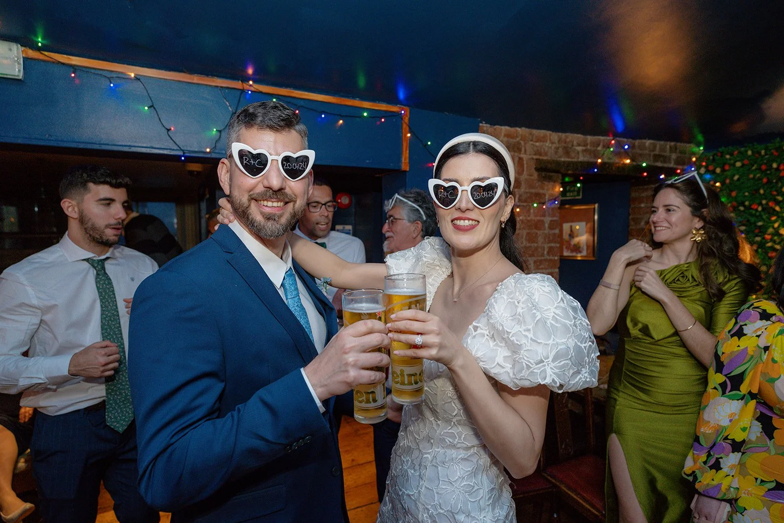 Ruth and Carmelo raise their drinks during their evening wedding reception at No. 4 Clifton Village in Bristol, UK, wearing playful heart-shaped sunglasses on the dance floor.