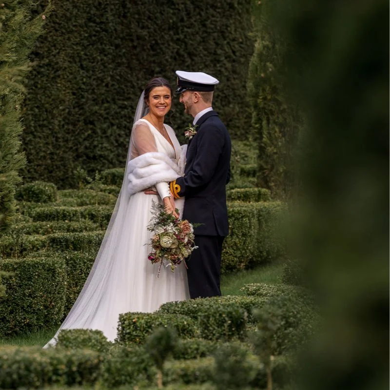 Megan & Luca stand together in the formal gardens at The Great Barn, Devon, framed by tall hedges and clipped greenery. This romantic wedding portrait captures the couple in soft light, with Luca in naval uniform and Megan holding her bouquet.