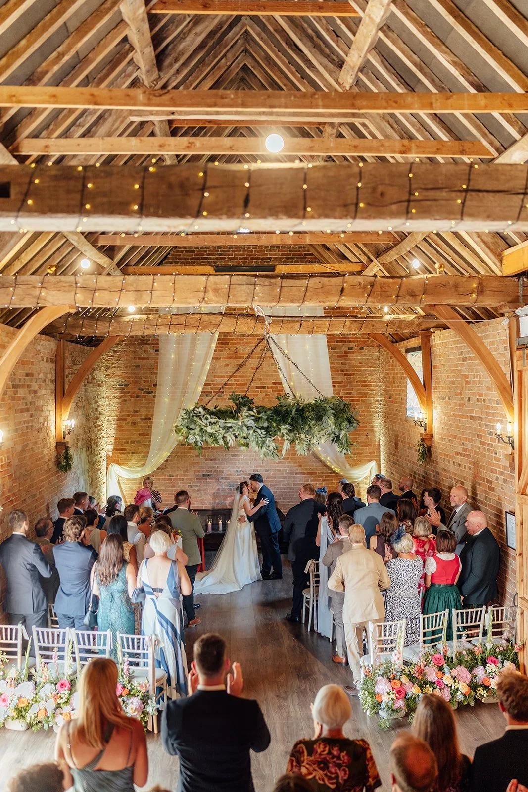 Harriet & Simon share their first kiss as guests applaud inside the rustic barn ceremony space at Dovecote Events in Oxfordshire, photographed by Just James Weddings.