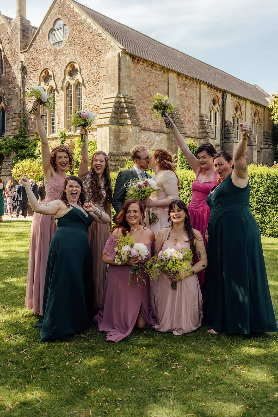 Charlotte & Jack wedding party portrait at Bishop’s Palace in Somerset, UK, with the couple kissing as bridesmaids cheer around them, raising bouquets on the sunny lawn beside the historic palace building during a joyful outdoor celebration.