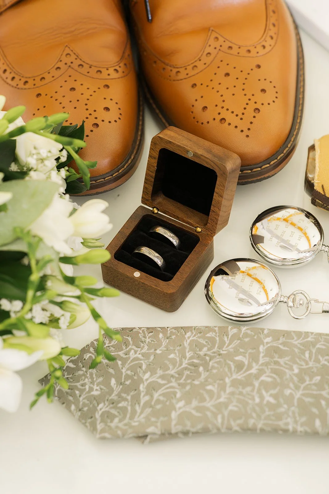 Wedding detail photo of Aaron & Jack’s rings in a wooden box beside tan brogue shoes, pocket watches, a patterned tie and white flowers at Rock Village Hall in Worcestershire, photographed by Just James Weddings.