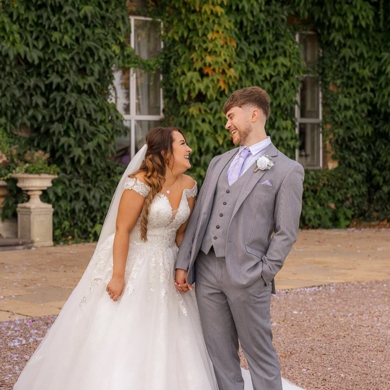 Rhiannon & Anthony laughing together while holding hands on their wedding day at Eastwood Park, gloucestershire. The bride’s lace gown and veil move softly as the groom smiles back at her, with ivy-covered stone walls behind them.