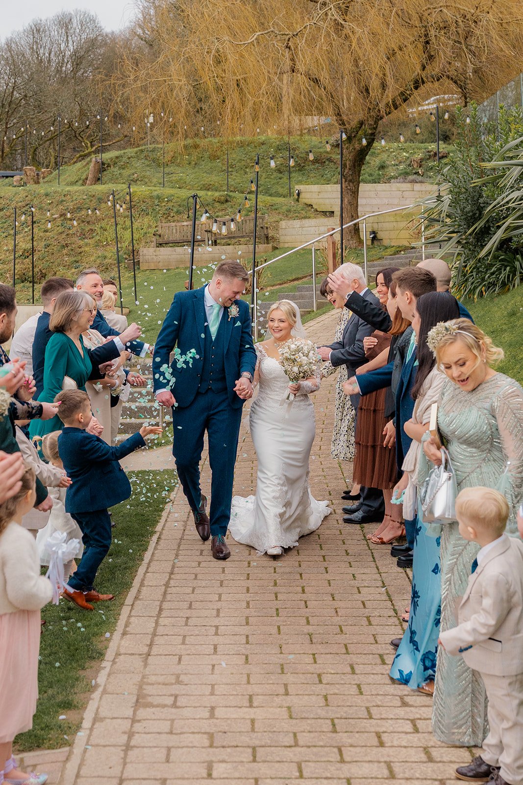 Andrew & Daisy walking hand in hand through a confetti line at Sandy Cove Hotel in Devon, surrounded by cheering guests and children, capturing joyful outdoor wedding celebrations against the coastal gardens of this popular seaside venue.
