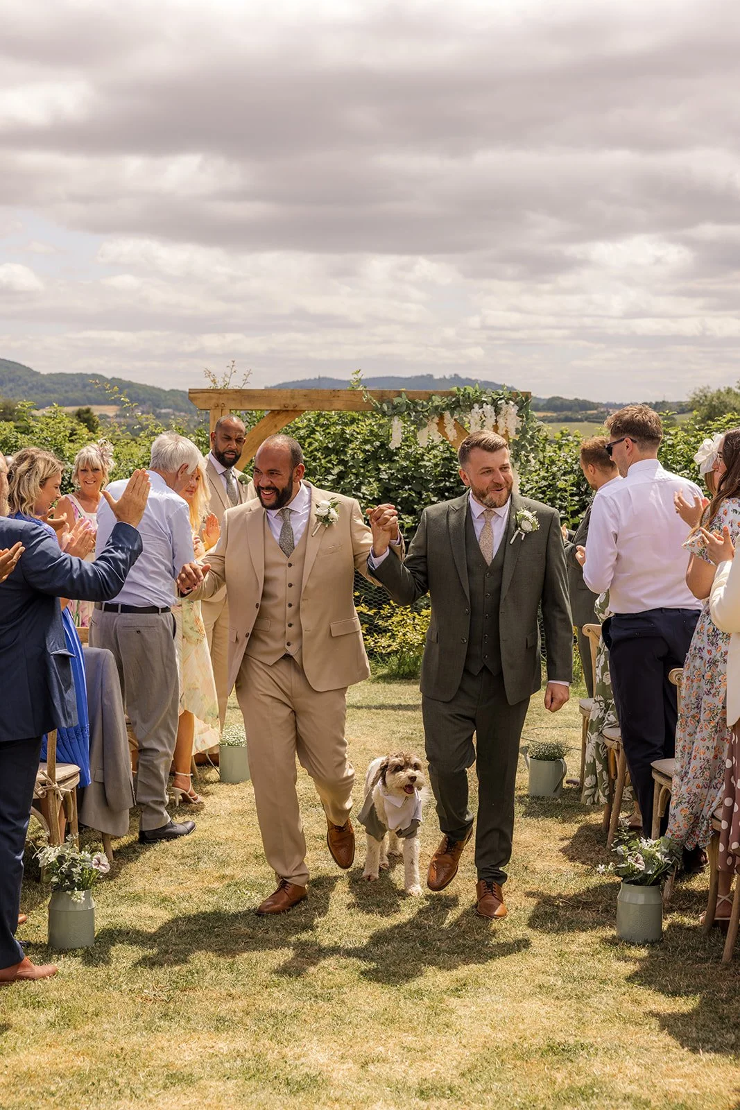Newlyweds Aaron & Jack walk hand in hand down the outdoor aisle with their dog Milo as guests applaud at Rock Village Hall in Worcestershire, photographed by Just James Weddings.