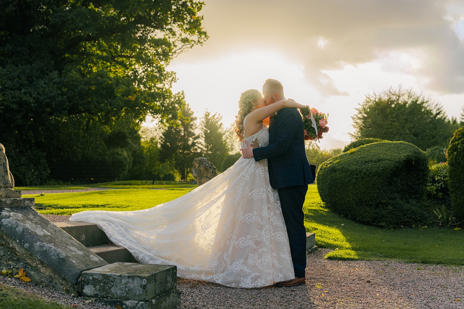 Lucy & Jay kissing in the gardens at Eastwood Park in Gloucestershire, with the bride’s veil glowing in warm evening light, photographed by Just James Weddings for romantic wedding portraits.