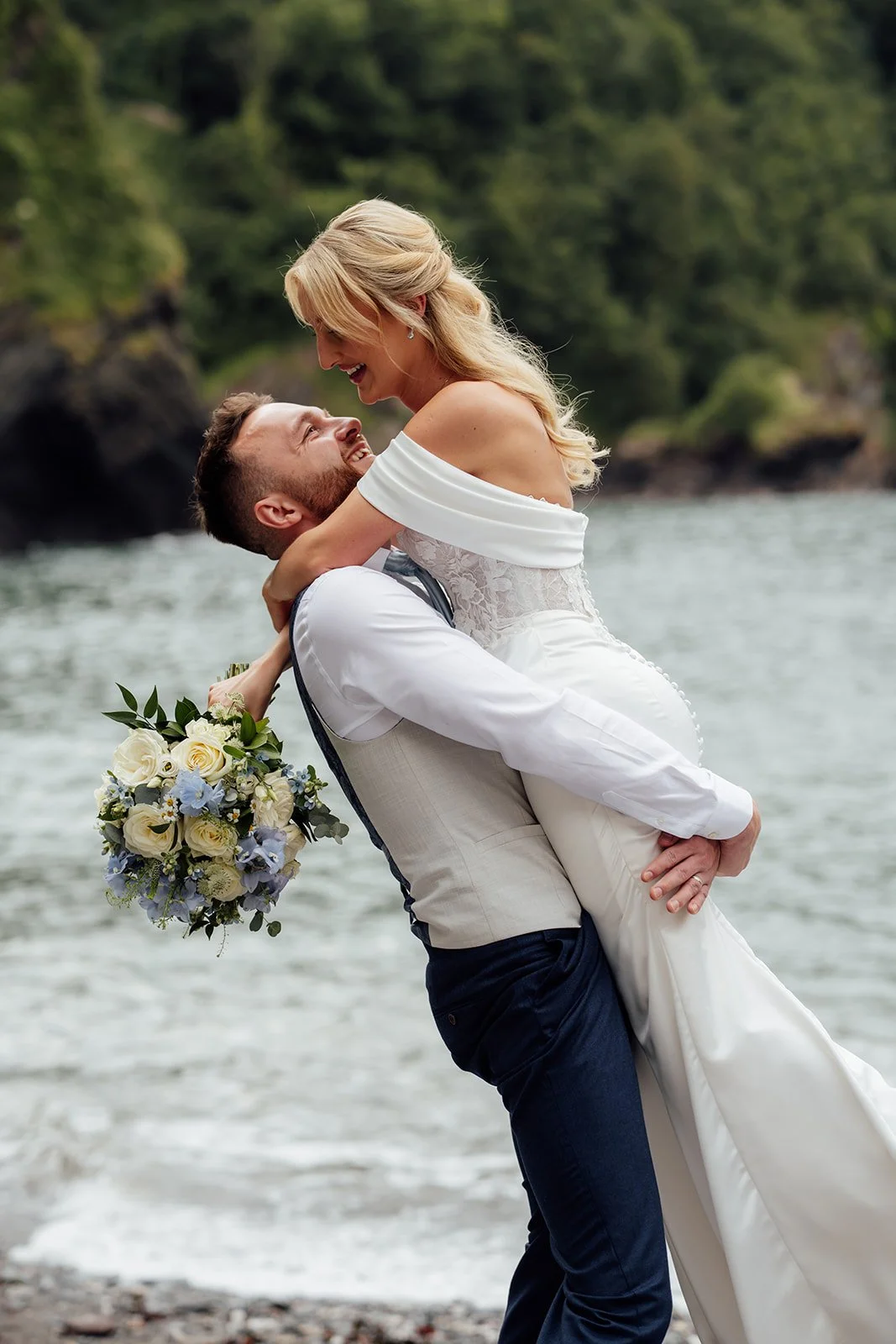 Amy & Connor laugh as the groom lifts the bride beside the sea at Sandy Cove Hotel in North Devon, UK, with her white and pale blue bouquet hanging at her side and wooded cliffs behind them.