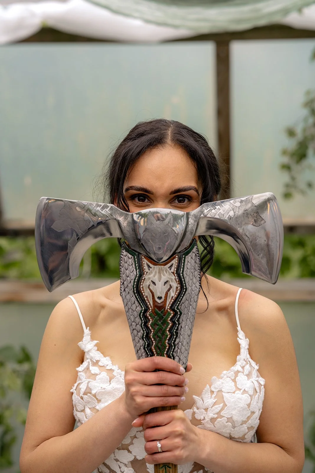 Jaye & Josh's Farrington's Farm wedding in Bristol with the bride holding a decorated double axe in front of her face, showing her eyes, lace wedding dress and ring in a playful portrait inside the greenery-filled poly tunnel ceremony space.