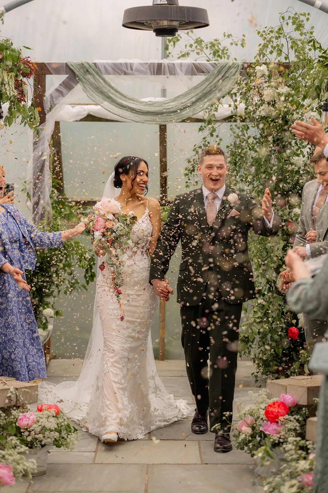 Jaye & Josh walk back down the aisle at Farrington's Farm in Bristol as confetti fills the poly tunnel, with guests cheering beside pink flowers, greenery, timber benches and their relaxed farm wedding ceremony setting full of pure joy and fun.