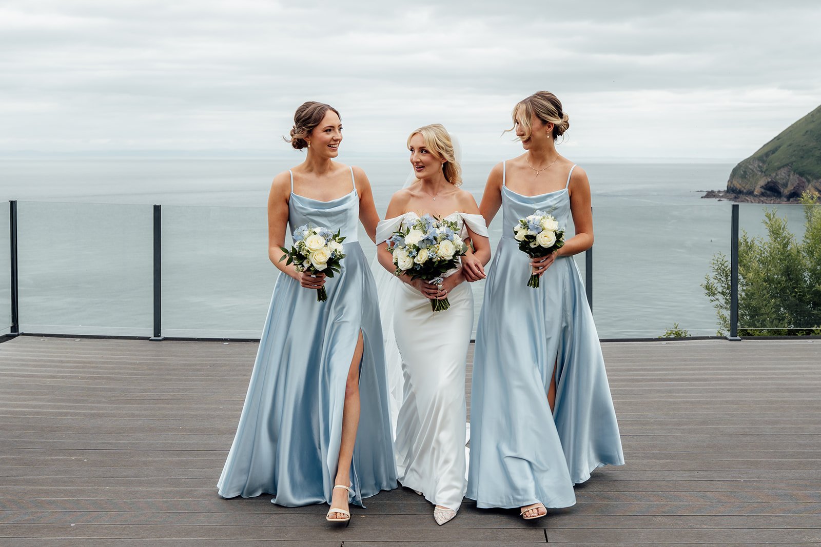 Amy walks between her two bridesmaids in pale blue dresses as they carry white and blue bouquets overlooking the sea at Sandy Cove Hotel in North Devon, UK.