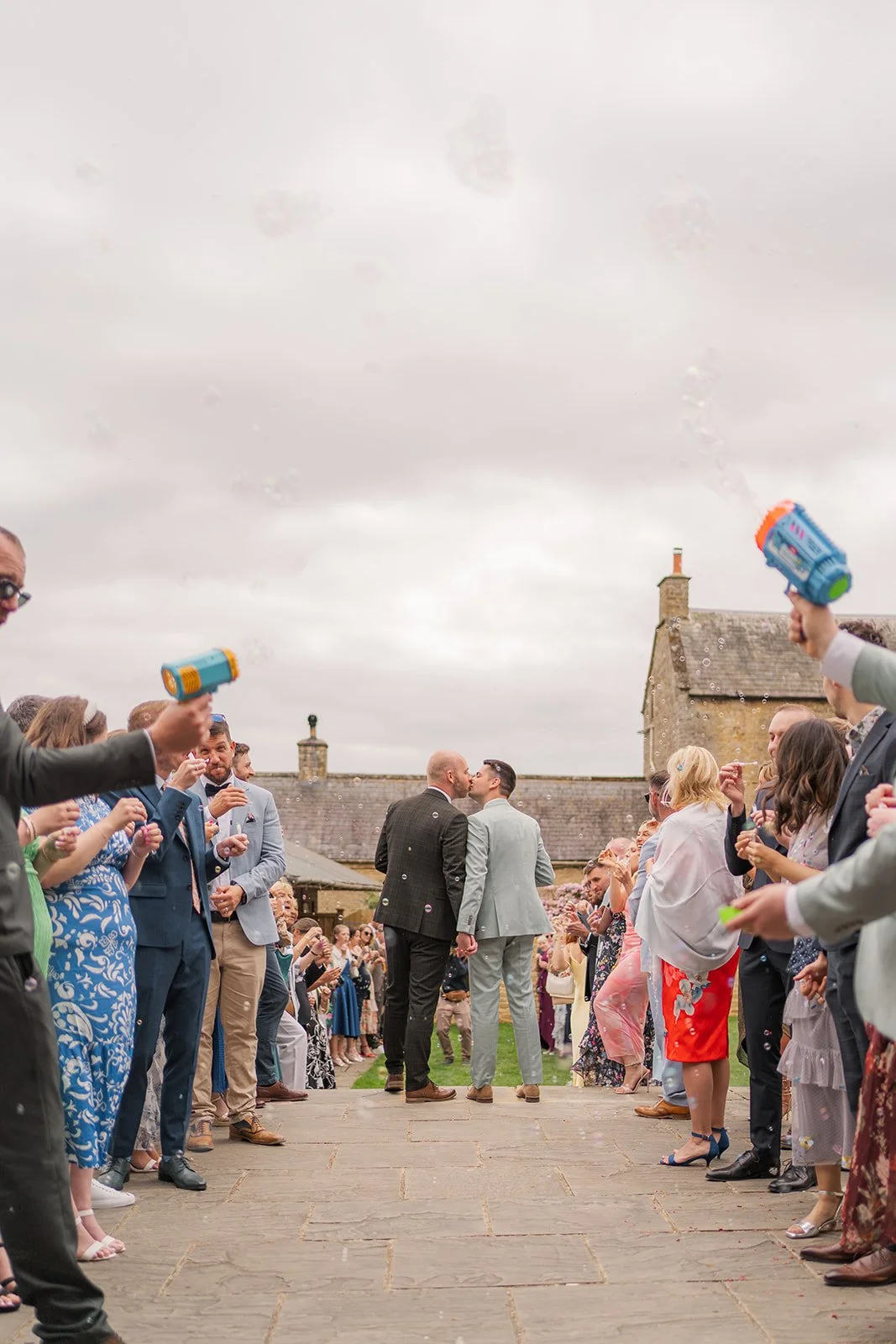 Gavin & Sam walk through a bubble-filled confetti exit at The Great Barn in Somerset, UK, surrounded by cheering wedding guests and colourful bubble guns, with the grooms sharing a kiss along the stone pathway outside the rustic barn venue.