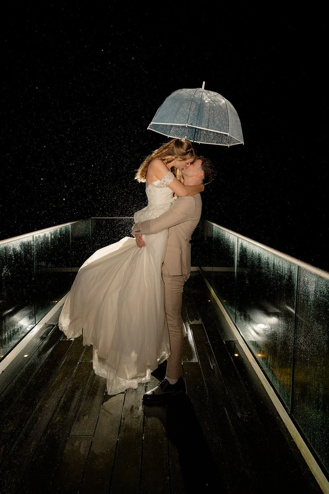 Hannah & Cameron share a romantic kiss beneath a clear umbrella on the terrace at Sandy Cove Hotel, North Devon. Rain falls around the couple as Cameron lifts Hannah in a dramatic nighttime portrait overlooking the coastal wedding venue.