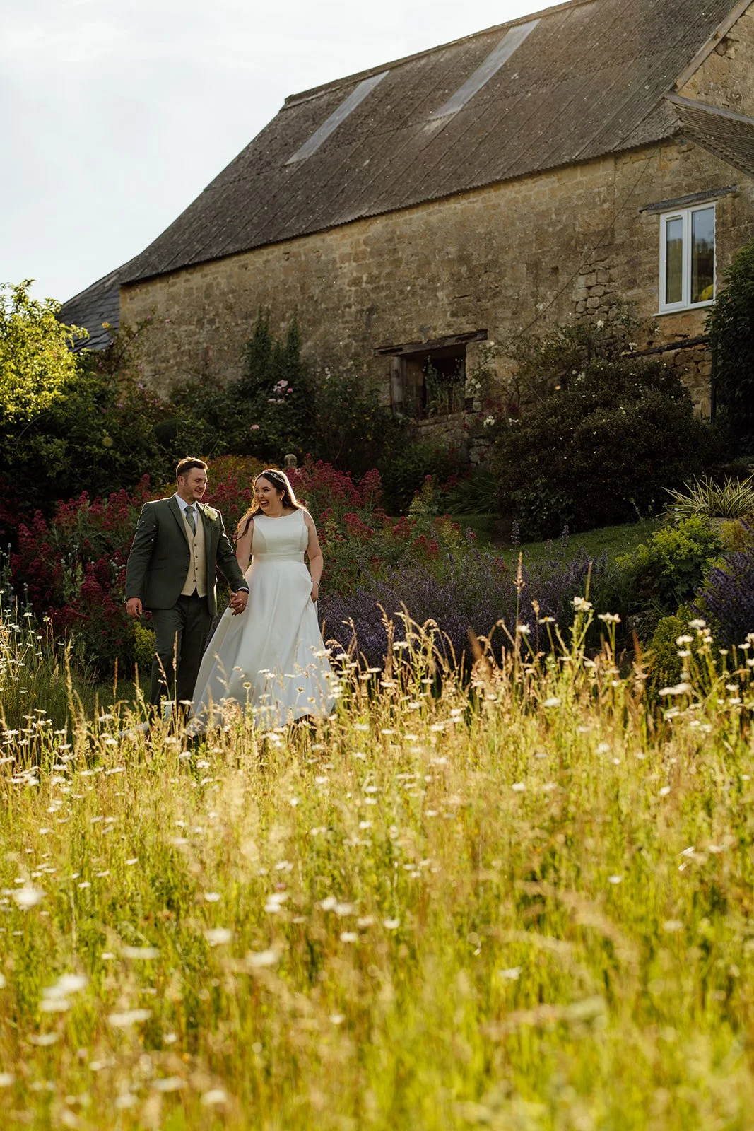 Chloe and Armani walk hand in hand through the wildflower gardens beside the stone barn at The Barn at Upcote in Gloucestershire, photographed by Just James Weddings.