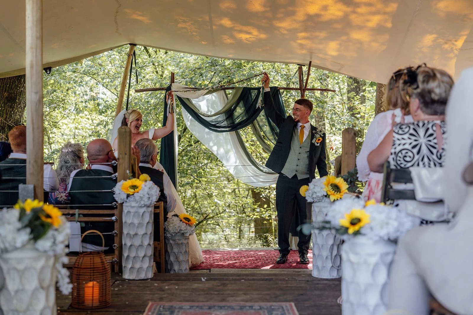 Lucy and Matthew raising their hands in celebration at the end of their woodland wedding ceremony at Curated Canvas at Noah’s Pond in Somerset, photographed by Just James Weddings.
