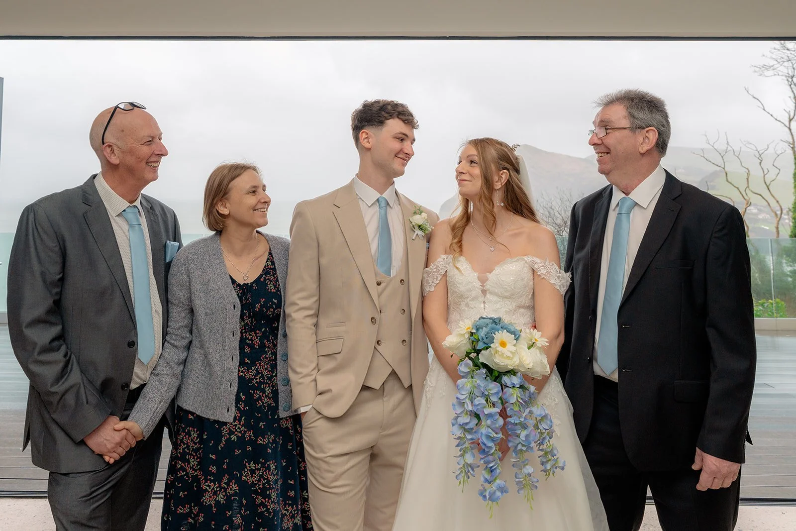 Hannah & Cameron stand with their parents during relaxed family portraits at Sandy Cove Hotel, North Devon. The couple smile at each other while holding a blue and white bouquet, with coastal views visible through the large windows behind them.