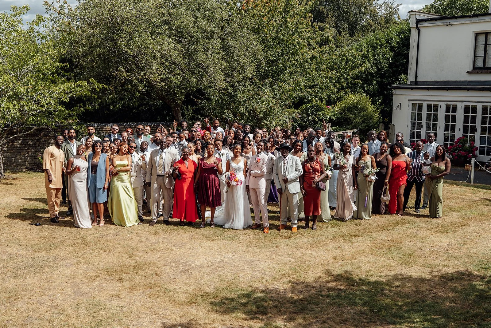 Kasey and Larisha standing with their wedding guests for a large group photo in the gardens at Oaks Farm Weddings in Surrey.