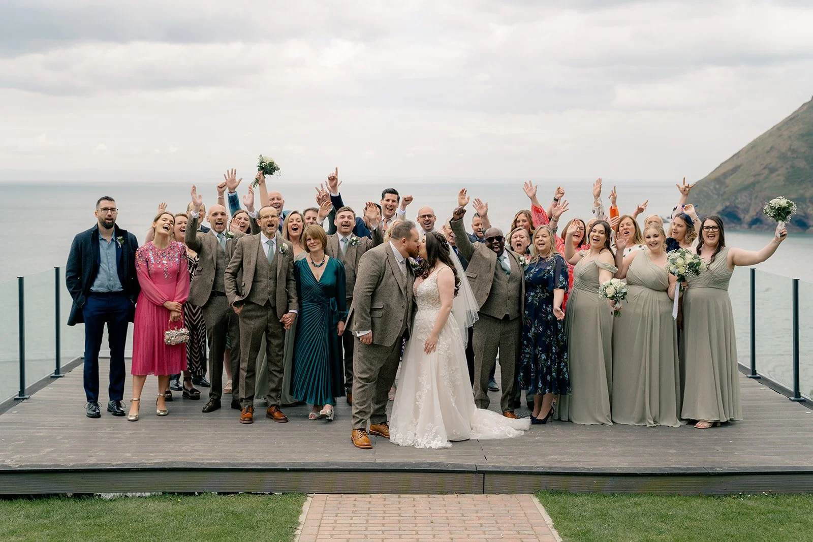 Sam & Terri celebrating with their wedding guests on the balcony at Sandy Cove Hotel in North Devon, with sea cliffs behind them and everyone cheering, photographed by Just James Weddings for fun coastal wedding group photos and inspiration.