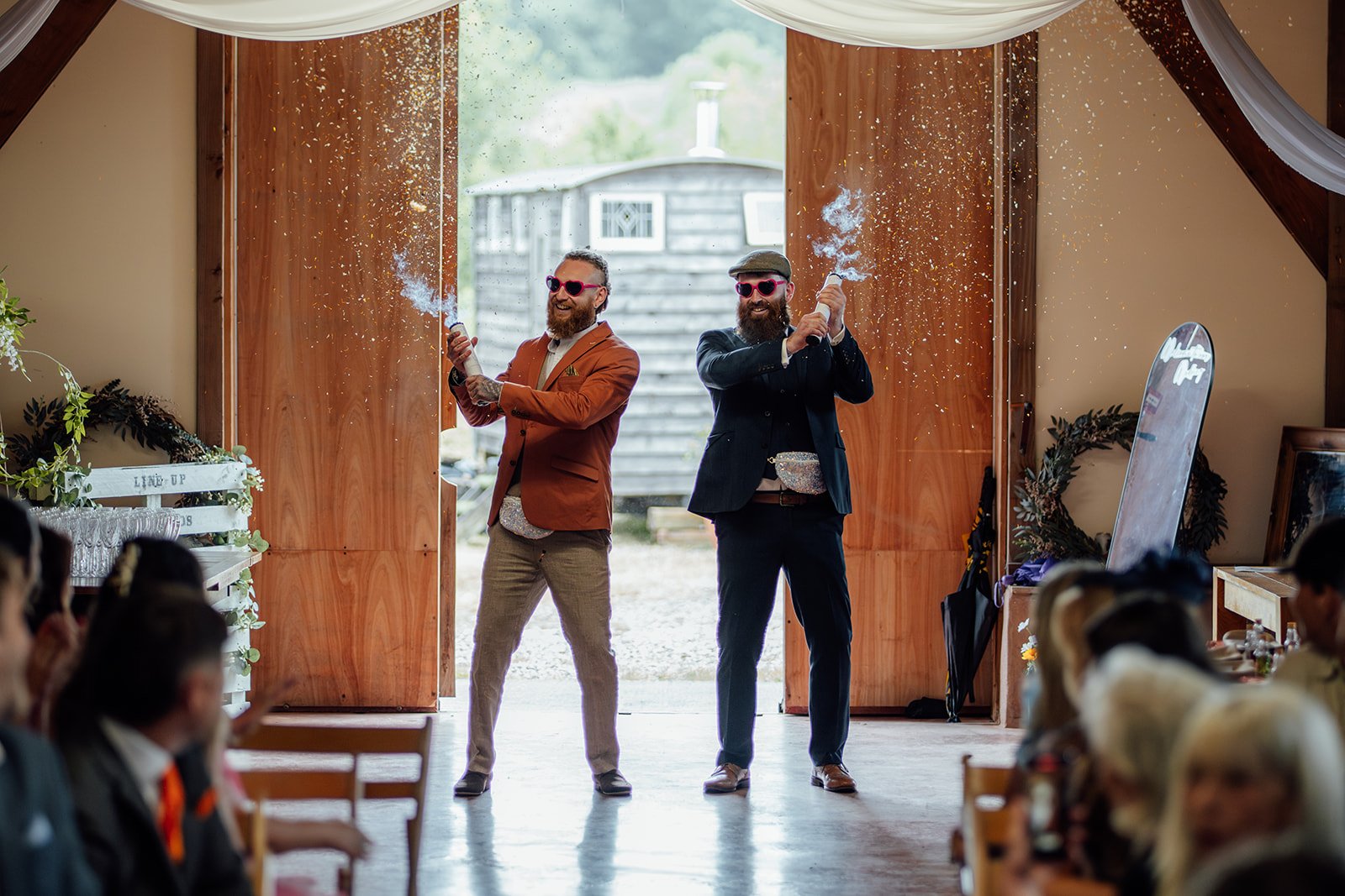 Fun ceremony entrance at Grays Barn in Devon, UK, with Ben and a groomsman wearing heart sunglasses and firing confetti cannons as guests look on inside the rustic barn, photographed candidly and colourfully by Just James Weddings.