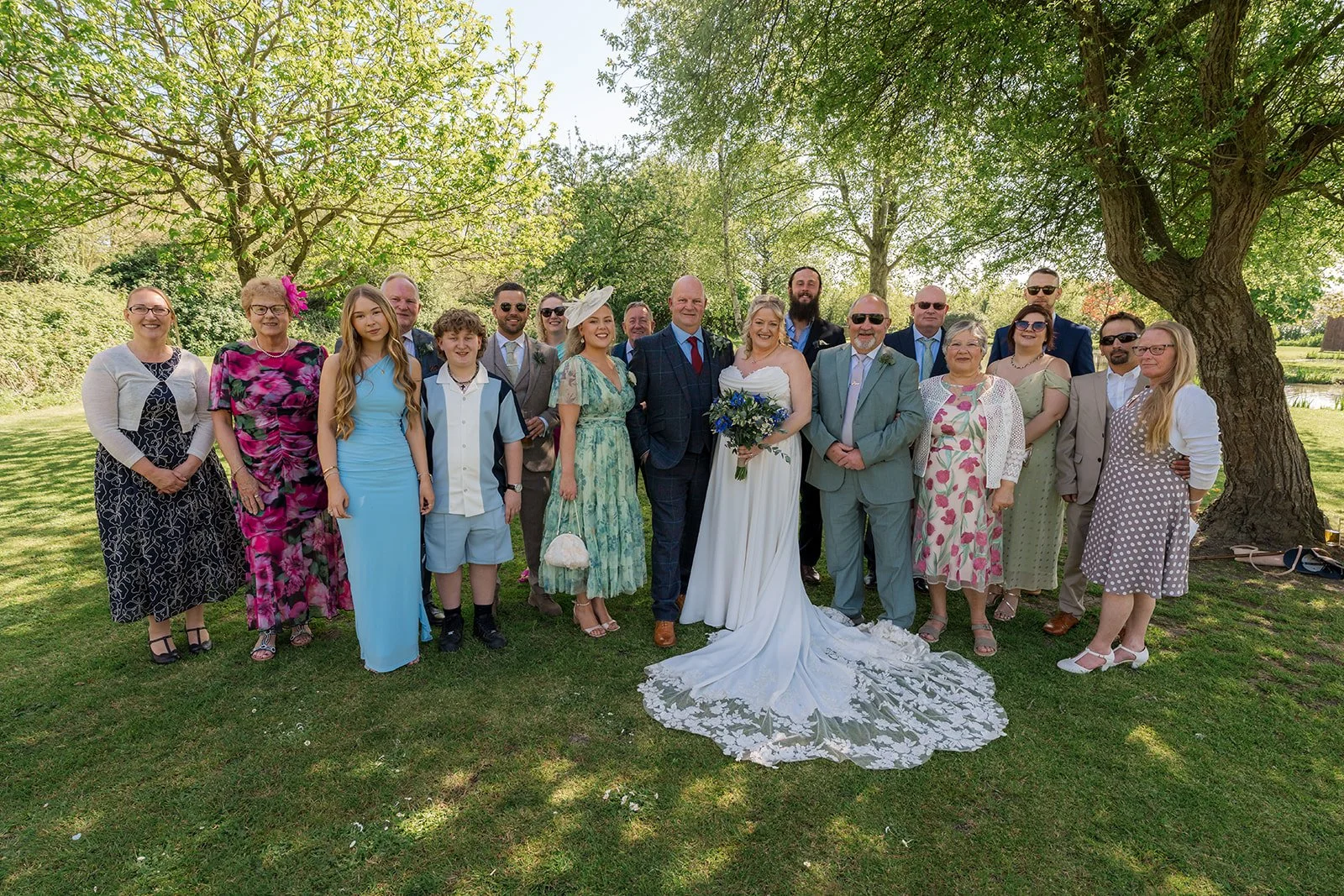 Debbie and Nigel stand with family and guests beneath the trees at Yarlington Barn in Somerset, creating a relaxed wedding group portrait with the lake nearby and Debbie’s lace train spread beautifully.